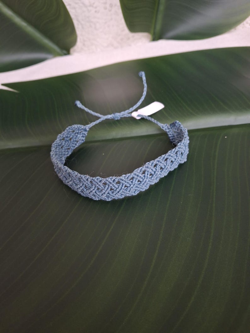 May include: A dusty blue macrame bracelet featuring an intricate woven chevron pattern. The bracelet is constructed from thin cord and includes adjustable tie closures with loose ends and a small white rectangular tag attached to one side.