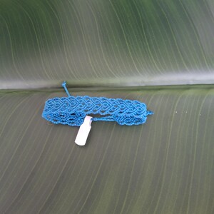 May include: A bright blue macrame bracelet featuring an intricate woven pattern of repeating diamond shapes. The bracelet is constructed from thin cord and includes a small, rectangular white paper tag attached to the center of the band.