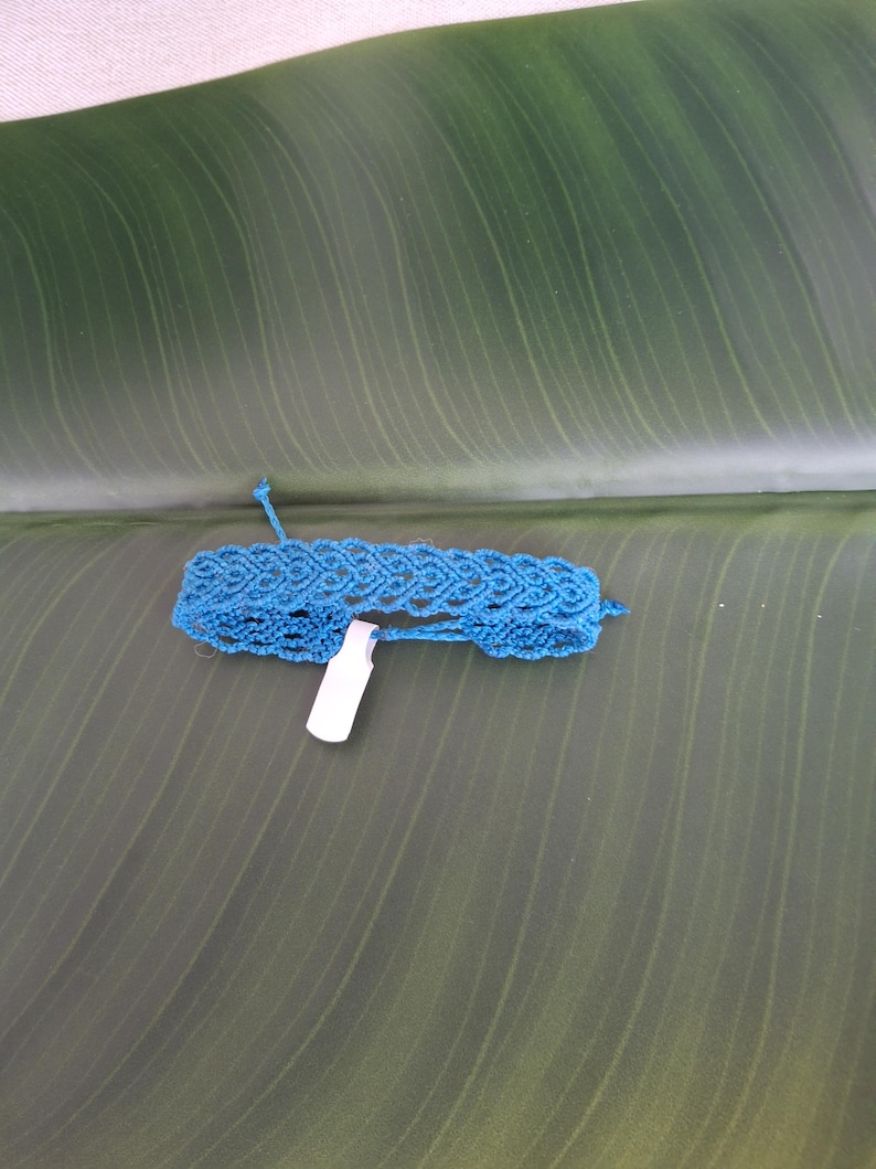 May include: A bright blue macrame bracelet featuring an intricate woven pattern of repeating diamond shapes. The bracelet is constructed from thin cord and includes a small, rectangular white paper tag attached to the center of the band.