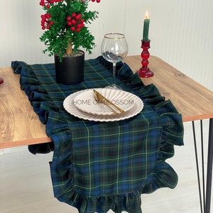 Könnte beinhalten: Ein gedeckter Tisch mit einem grün-blau karierten Tischläufer mit Rüschenkante. Ein weißer Teller mit goldenem Besteck liegt auf dem Läufer. Eine kleine Topfpflanze mit roten Beeren und ein Weinglas stehen ebenfalls auf dem Tisch.