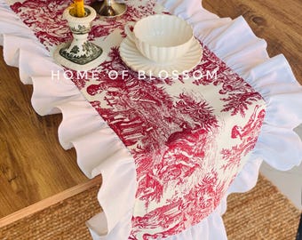 Può includere: Una tovaglia in toile de Jouy rossa e bianca con un bordo bianco a balze. La tovaglia presenta una scena pastorale dettagliata. Una candela accesa è in un portacandele decorativo e una tazza e un piattino sono sulla tovaglia. Il testo "HOME OF BLOSSOM" è visibile.