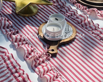 May include: A festive table setting featuring a red and white striped table runner with ruffled edges. A gold star-shaped tray and two white candles with heart details are on the table. The text "HOME OF BLOSSOM" is visible.