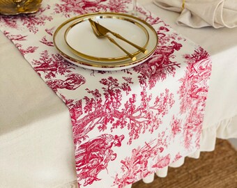 Può includere: Una tovaglia bianca con un motivo toile de Jouy rosso raffigurante una scena pastorale. La tovaglia è posizionata su una tovaglia bianca, con piatti, posate e un bicchiere da vino bordati d'oro. Una candela e un tovagliolo completano l'elegante mise en place.