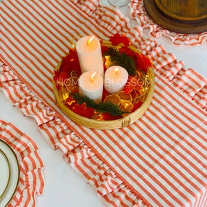 May include: A table setting with a red and white striped table runner with frilled edges. A round woven tray holds three white candles, greenery, and dried citrus. The text "HOME OF BLOSSOM" is visible. Matching placemats and a plate are also present.