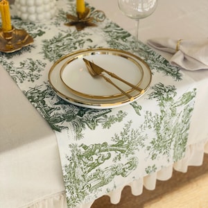 Könnte beinhalten: Eine elegante Tischdekoration mit weißer Tischdecke, grün-weißem Toile-de-Jouy-Tischläufer, goldgeränderten Tellern und passendem Besteck. Ein Weinglas und Kerzen ergänzen das Arrangement.