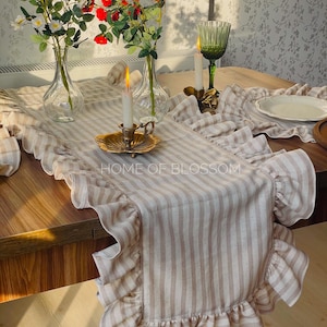 May include: A beige and white striped table runner with ruffled edges. The text "HOME OF BLOSSOM" is printed on the runner. The table is set with flowers in vases, candles, and a green glass.