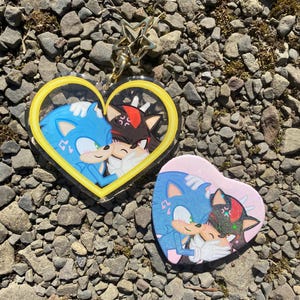 May include: Two heart-shaped charms featuring Sonic the Hedgehog and Shadow the Hedgehog. The charms are made of clear acrylic with a yellow outline and a pink outline. The charms are on a gray gravel background.