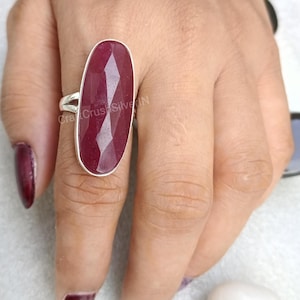 May include: A silver ring with a large, oval-shaped, faceted, dark red gemstone. The ring is on a person's finger.