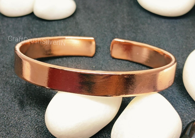 Peut inclure: Un bracelet en cuivre brillant avec une finition lisse et polie. Le bracelet est ouvert aux deux extr&eacute;mit&eacute;s et pr&eacute;sente un design simple et minimaliste.