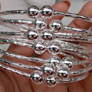 May include: A set of six silver bangles with textured surfaces and round silver beads. The bangles are stacked on top of each other.