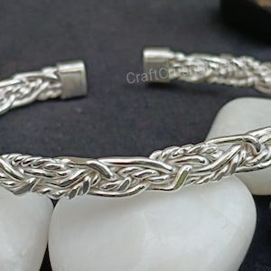 Puede incluir: Un brazalete plateado con un diseño trenzado. El brazalete está abierto en los extremos y tiene un cierre de broche. El brazalete se exhibe sobre piedras blancas sobre un fondo oscuro.