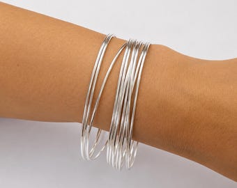 May include: A set of multiple thin, polished silver-colored bangle bracelets worn together. The bracelets have a smooth, reflective metallic finish and a simple, circular design that sits stacked around the wrist area.