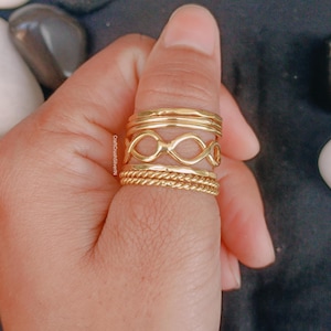 May include: A collection of gold-colored rings in various designs, including infinity symbols and twisted rope patterns. The rings are stacked on a finger, showcasing their different textures and styles. The rings are made of a shiny, metallic material.