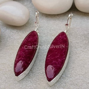 May include: A pair of dangle earrings featuring elongated, oval-shaped, deep red stones set in silver-colored metal. The earrings have a lever-back closure. The text "CraftCrushSilverIN" is visible on the earrings.