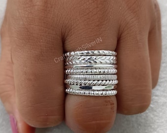 May include: A stack of silver rings with different textures and patterns. The rings are worn on a person's finger.