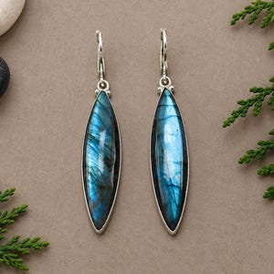 Puede incluir: Un par de pendientes largos en forma de marquesa con piedras preciosas de labradorita pulida con destellos azules iridiscentes. Las piedras están engastadas en marcos de metal plateado y unidas a cierres de tipo dormilona en metal plateado.