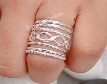 May include: A set of seven silver rings stacked on a finger. The rings are different styles, including a twisted rope design, a simple band, and an infinity symbol.