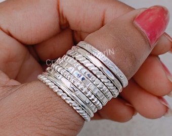 May include: A stack of silver rings with different textures and patterns. The rings are worn on a person's finger.