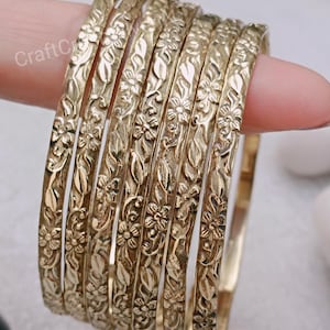 May include: Set of seven gold-toned metal bangles with a floral design. The bangles are stacked on top of each other.