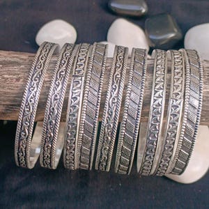 May include: A set of silver bangles with detailed patterns. The bangles are presented on a wooden piece, with white and black stones in the background. Each bangle displays a unique design, demonstrating fine craftsmanship.