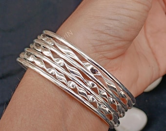 Pode incluir: Um conjunto de seis pulseiras de prata com um design torcido. As pulseiras estão empilhadas no pulso de uma pessoa.