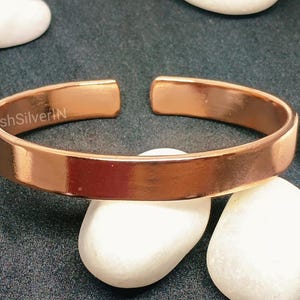 Peut inclure: Un bracelet en cuivre brillant avec une finition lisse et polie. Le bracelet est ouvert aux deux extr&eacute;mit&eacute;s et pr&eacute;sente un design simple et minimaliste.