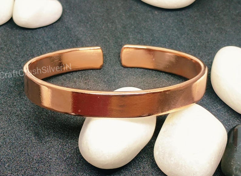 Peut inclure: Un bracelet en cuivre brillant avec une finition lisse et polie. Le bracelet est ouvert aux deux extr&eacute;mit&eacute;s et pr&eacute;sente un design simple et minimaliste.