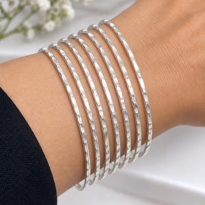 Puede incluir: Un conjunto de siete brazaletes finos de color plateado con una superficie martillada y texturizada. Cada brazalete circular tiene un acabado metálico reflectante y se lleva apilado en la muñeca, creando una apariencia uniforme y superpuesta.