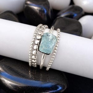 May include: A silver ring featuring a rectangular, raw, light blue aquamarine stone set in the centre. The band is composed of three distinct strands: two outer beaded silver chains and a smooth, solid silver centre band holding the stone.