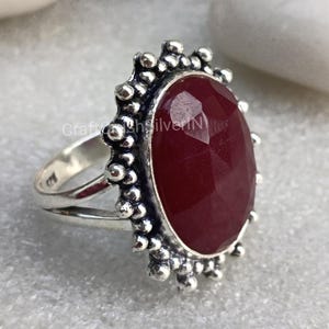 Puede incluir: Un anillo de plata con una piedra preciosa grande, ovalada, de color rojo oscuro engastada en un engaste decorativo con cuentas. El anillo tiene una banda sencilla.