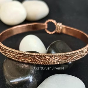 Puede incluir: Un brazalete de color cobre con un intrincado diseño floral y de hojas. El brazalete tiene un cierre de broche y se exhibe sobre piedras blancas y negras. El nombre de la tienda Etsy "CraftCrushSilverIN" es visible.
