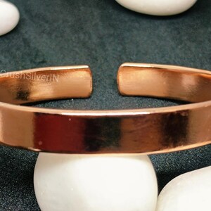 Peut inclure: Un bracelet en cuivre brillant avec une finition lisse et polie. Le bracelet est ouvert aux deux extr&eacute;mit&eacute;s et pr&eacute;sente un design simple et minimaliste.