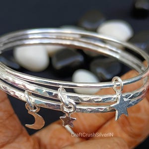 May include: Three silver bangles with hammered texture and dangling charms. Charms include a crescent moon and two stars. The bangles are displayed against a dark background with white and black stones.