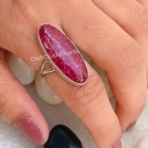 May include: A silver ring with a large, oval-shaped, pink gemstone. The ring is on a finger and the stone has a speckled pattern.