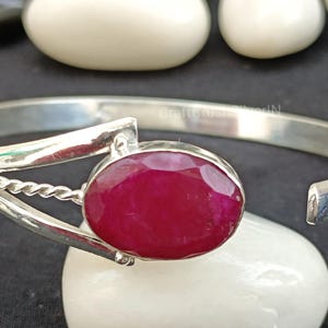 Op de afbeelding: Een zilveren armband met een grote, gefacetteerde, roze edelsteen in het midden. De armband heeft aan de ene kant een gedraaid draadontwerp.