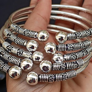 May include: A set of six silver bangles with intricate designs and round accents. The bangles are stacked on top of each other and have a textured finish.