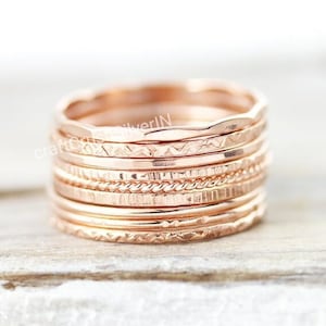 Può includere: Un set di nove anelli impilabili in oro rosa con diverse trame e motivi. Gli anelli sono impilati uno sopra l'altro e disposti in cerchio.