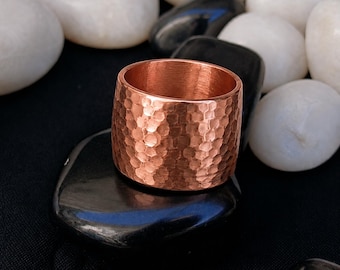 Può includere: Un anello a fascia larga realizzato in rame lucido, caratterizzato da una distinta texture martellata su tutta la superficie esterna. Il metallo presenta una calda tonalità metallica bruno-rossastra con riflessi sulla superficie.