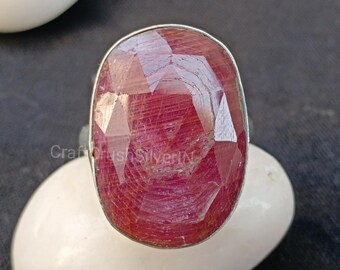 May include: A silver ring with a large, faceted, oval-shaped ruby gemstone. The ruby is a deep red color with a slight pink hue.