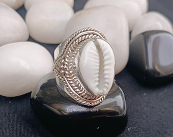 May include: A silver ring featuring a white seashell centerpiece. The ring has a detailed, textured band with a braided design and floral accents. The ring is displayed on a black stone, surrounded by white and black pebbles.