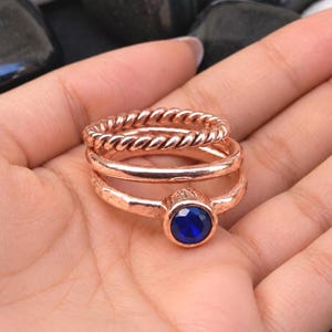May include: A set of three stacked rings in a rose gold tone. The top ring features a twisted rope texture, the middle ring has a smooth polished band, and the bottom ring is a hammered band set with a single round, deep blue faceted stone.