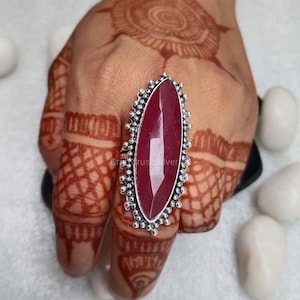 May include: A silver ring with a large, oval-shaped, dark red gemstone. The ring has a decorative, beaded border.