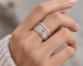 May include: A set of five stacked silver rings worn on a finger. The rings feature varied textures, including smooth polished bands, a beaded design, and a subtle hammered finish, creating a layered metallic appearance across the finger joint.