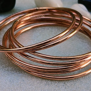 Puede incluir: Un conjunto de siete brazaletes de cobre, apilados uno encima del otro. Los brazaletes son lisos y brillantes, de un cálido color cobre.