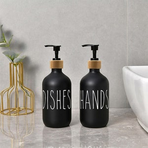 May include: Two black glass soap dispensers with wooden tops and white lettering. The dispenser on the left says "DISHES" and the dispenser on the right says "HANDS".