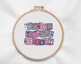 Könnte beinhalten: Ein Kreuzstich-Stickrahmen mit einem rosa, violetten und blauen Design, das "NOT YOUR BABY" in Blockbuchstaben zeigt. Die Buchstaben sind versetzt angeordnet, um wie Spielzeugblöcke auszusehen.