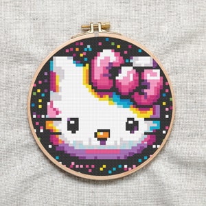 以下が含まれることがあります： Hello Kittyのピクセルアートが刺繍されたクロスステッチの刺繍枠。虹色のカラーリングで、Hello Kittyの頭にはピンクのリボンが飾られています。背景は黒で、カラフルなコンフェッティが散りばめられています。