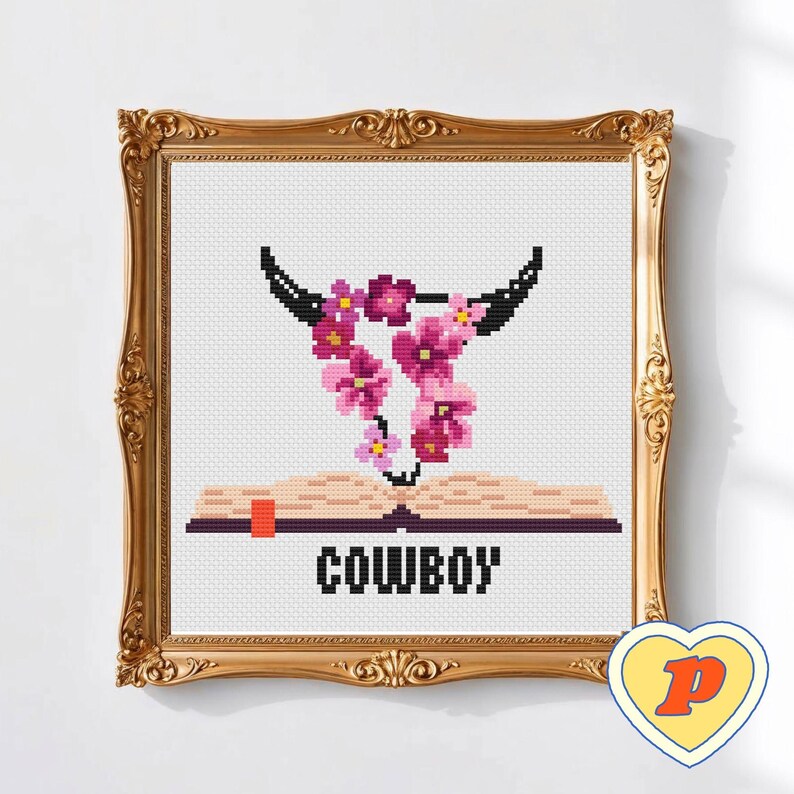 May include: Cross-stitch artwork in a gold frame. The design shows a bull skull with pink flowers, above an open book with the word "COWBOY". The background is white.
