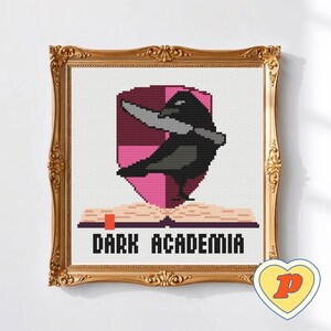 May include: Cross-stitch artwork in a gold ornate frame. The design shows a black crow with a knife, set against a pink shield, above an open book. The text "DARK ACADEMIA" is below the book.