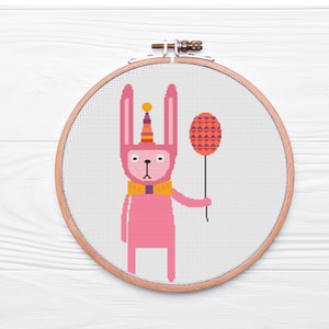 May include: Pink bunny rabbit wearing a party hat and holding a pink and purple balloon. The bunny is embroidered on a white fabric in a round wooden embroidery hoop.
